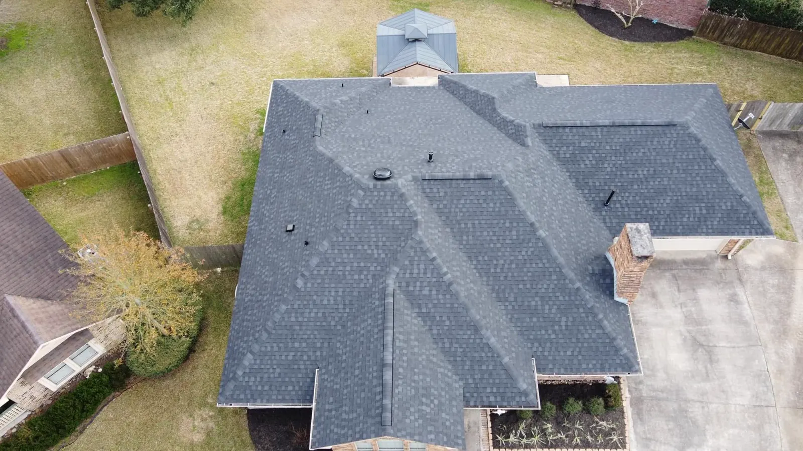 Drone view straight down onto a charcoal shingle roof, a fenced backyard and a driveway.