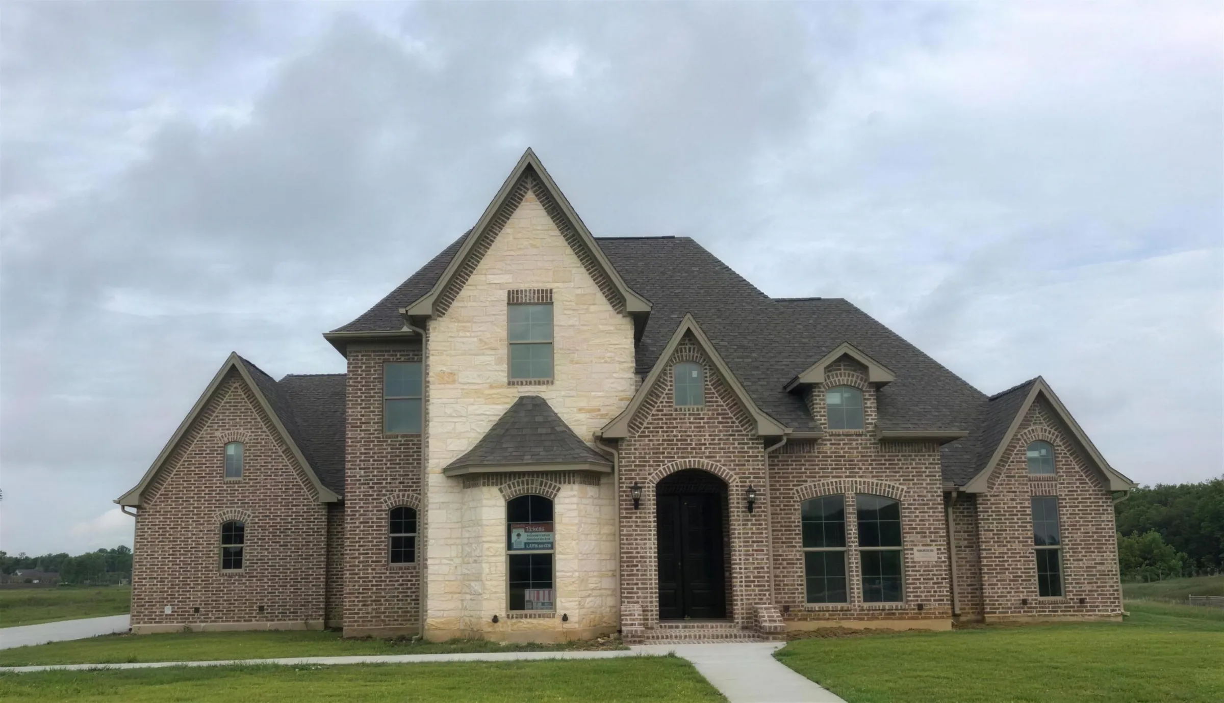 A two-storey brick and limestone house with a dark shingle roof under a grey Southeast Texas sky, from ABCO's job photos.